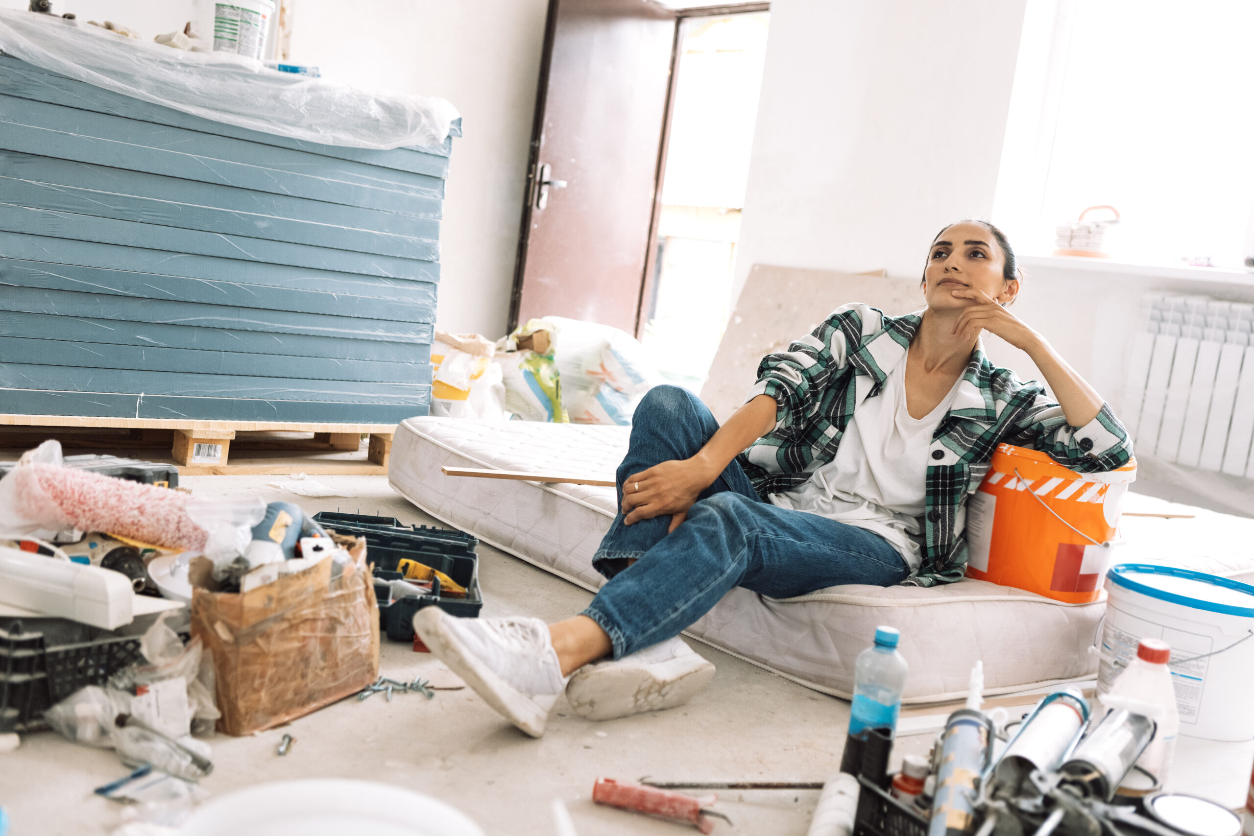 a young woman is sitting on a mattress among building materials and inventory. a charming girl in casual clothes is thinking about home renovation.